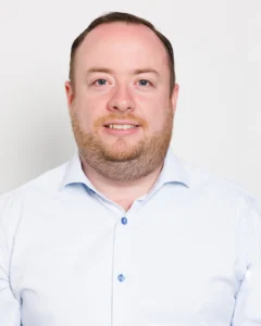 Headshot_GaryKing_01 Head-and-shoulders portrait of a smiling man in a light blue button-down shirt against a white background