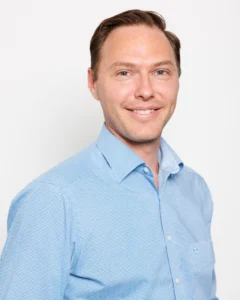 Headshot_Jake_01 Portrait of a smiling man in a light blue checkered shirt against a white studio background.