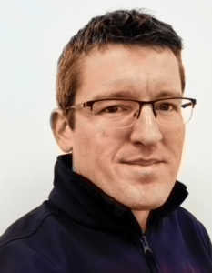STEPHEN-HEADHSOT-WITH-COLOUR-min Portrait of a man with short dark hair and rectangular glasses, wearing a dark zip-up jacket, looking at the camera.